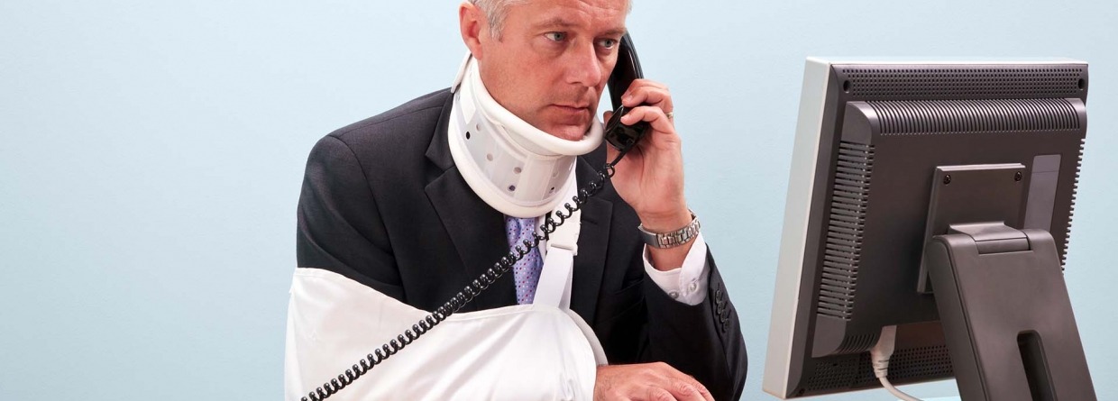 Gewonde zakenman met arm in mitella en nekbrace zit achter zijn computer aan zijn bureau aan de telefoon